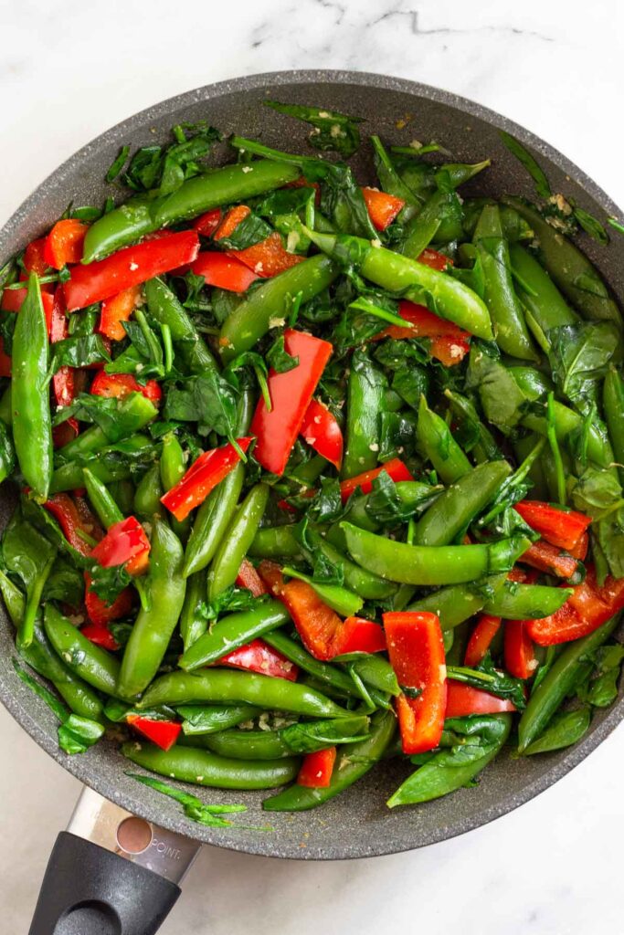 Skillet filled with sautéed snap peas, sliced bell peppers, and spinach.