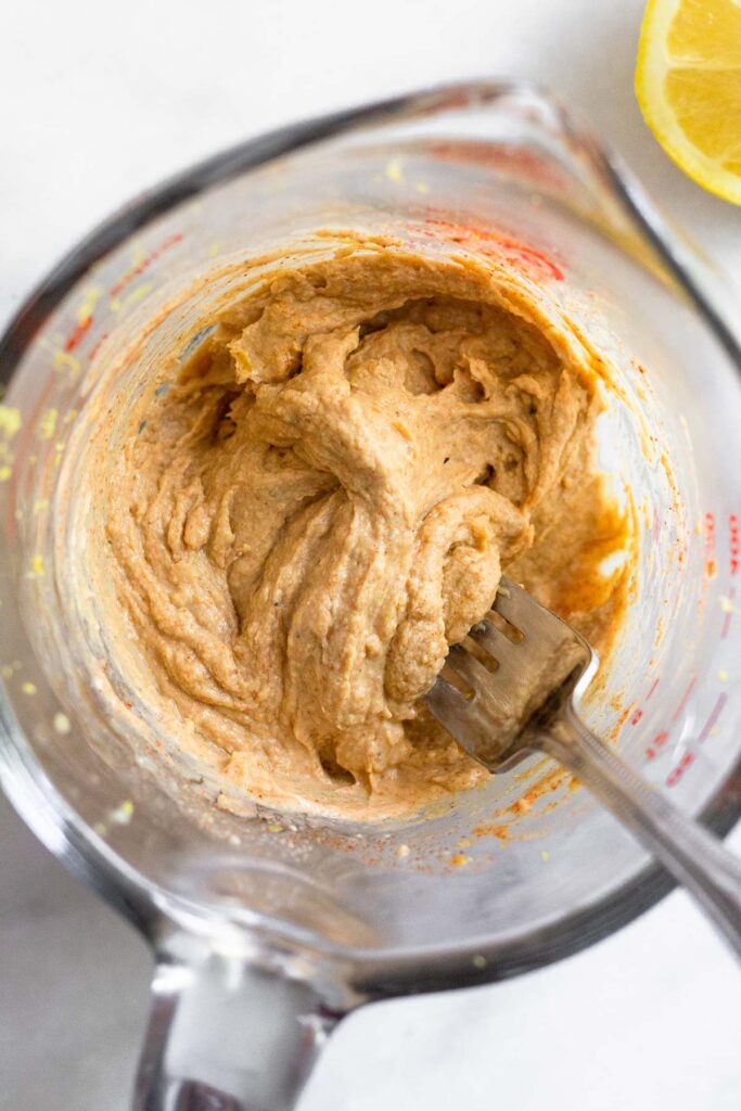 A mixture of tahini a spices in a large measuring sup with a fork in it. Next to the cup is half a lemon.