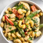 A bowl of tahini pasta with chicken and veggies. It is garnished with fresh herbs. Around the bowl is half a lemon, a ramekin of spices, and a pan.