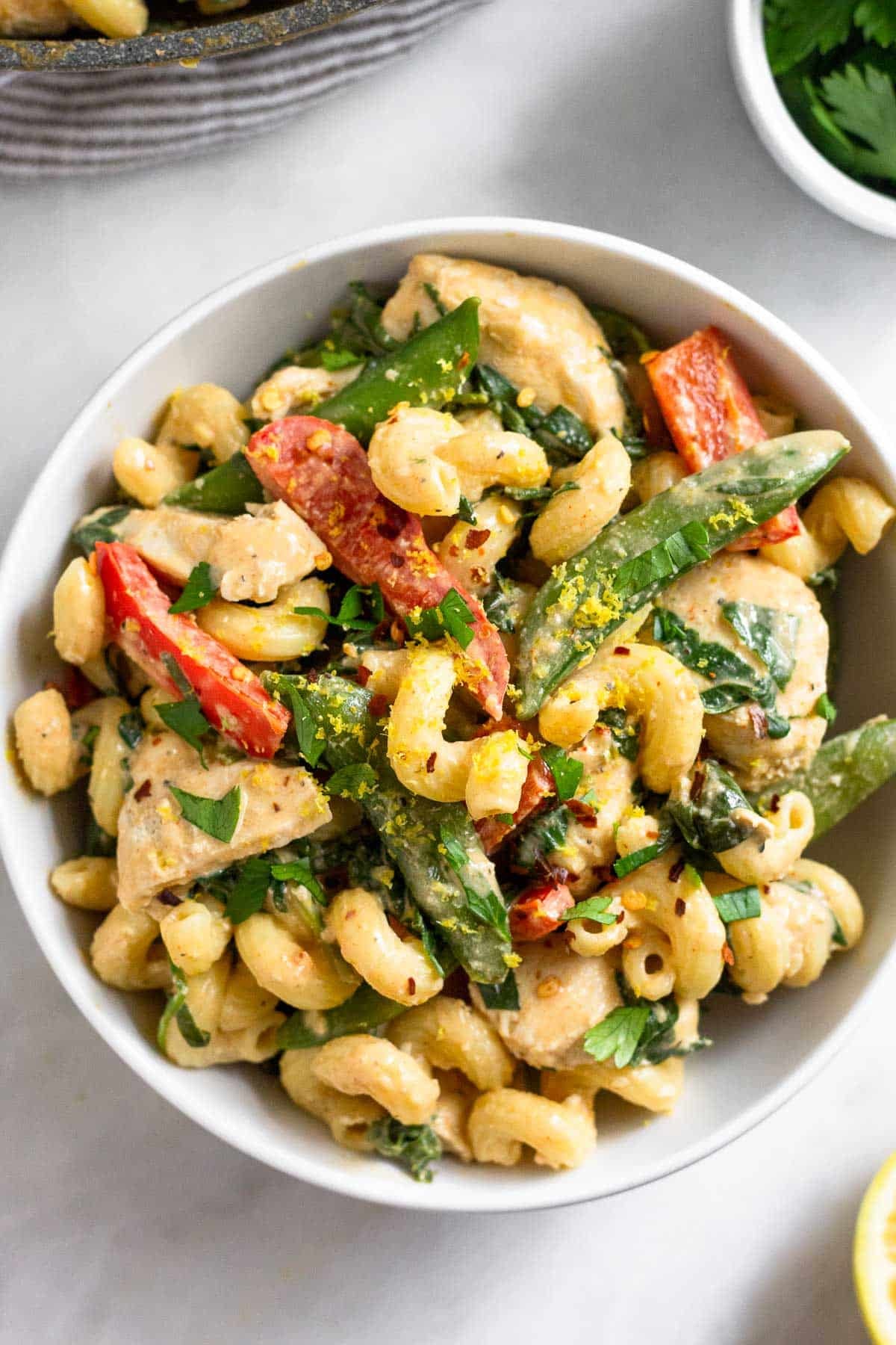 A bowl of tahini pasta with chicken and veggies. It is garnished with fresh herbs. Around the bowl is half a lemon, a ramekin of spices, and a pan.