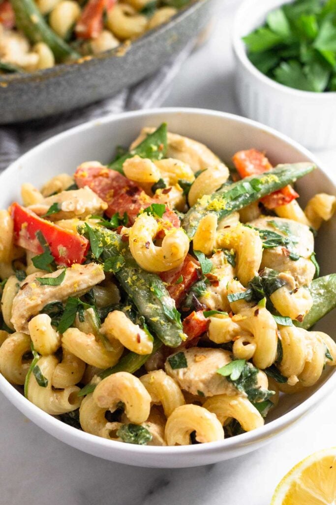 Bowl of chicken tahini pasta with veggies topped with herbs and lemon zest. Around the bowl is half a lemon, a dish of more fresh herbs, and a pan of more pasta.