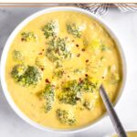 Bowl of vegan broccoli cheddar soup with a spoon coming out of it and topped with red pepper flakes. Behind it is a striped bowl and in front of it is another bowl of soup.
