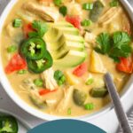 Overhead shot of cheesy chicken soup in a bowl topped with avocado, jalapeños, and herbs. A spoon is in the bowl and around it is a plate with more herbs, an avocado, a bowl of chips, and a kitchen towel.