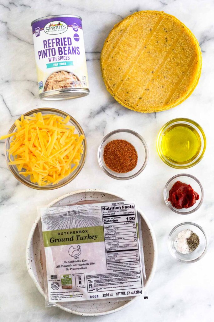 Marble counter with a can of refried beans, a stack of corn tortillas, a bowl of olive oil, a bowl of tomato paste, a bowl of salt and pepper, a bowl of taco seasoning, a package of ground turkey, and a bowl of shredded cheese.