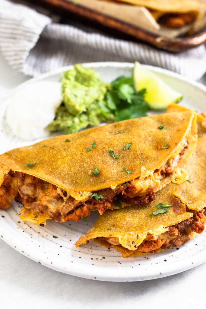 Two crispy tacos on a plate garnished with cilantro. Also on the plate is a lime wedge, sliced jalapeño, guacamole, and sour cream.