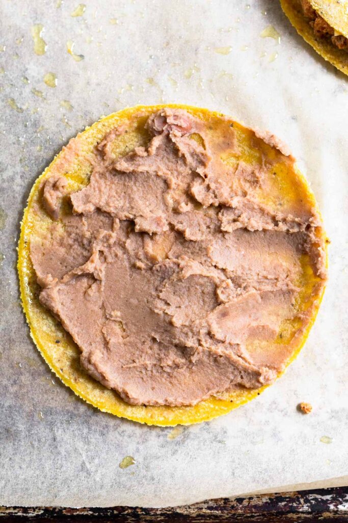 Corn tortilla with refried beans spread across it on a baking sheet lined with parchment paper.