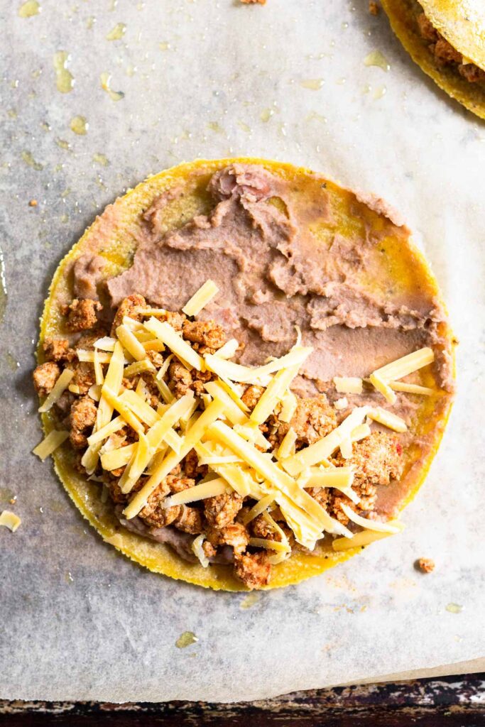 Corn tortilla with refried beans spread across it. Half of it has ground turkey and shredded cheese on it. It is on a baking sheet lined with parchment paper.