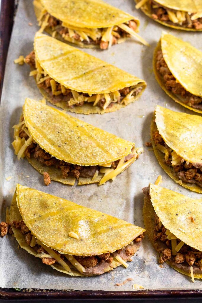 Corn tortillas that are folded in half and stuffed with beans, meat, and cheese on a baking sheet.