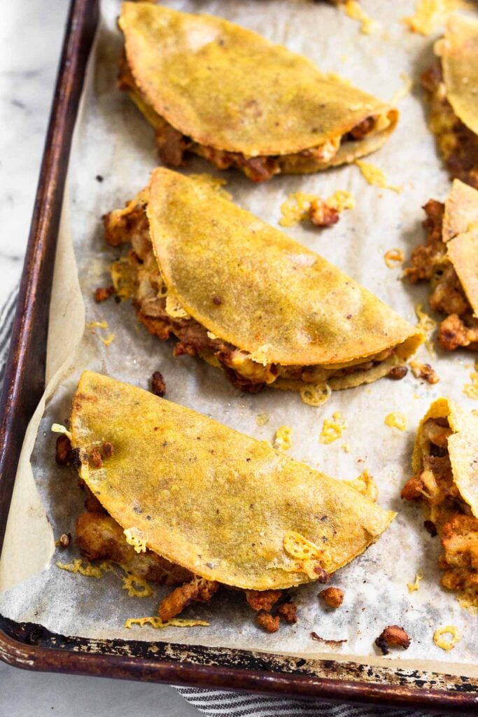 Crispy tacos on baking sheet with a little meat and cheese coming out of them.