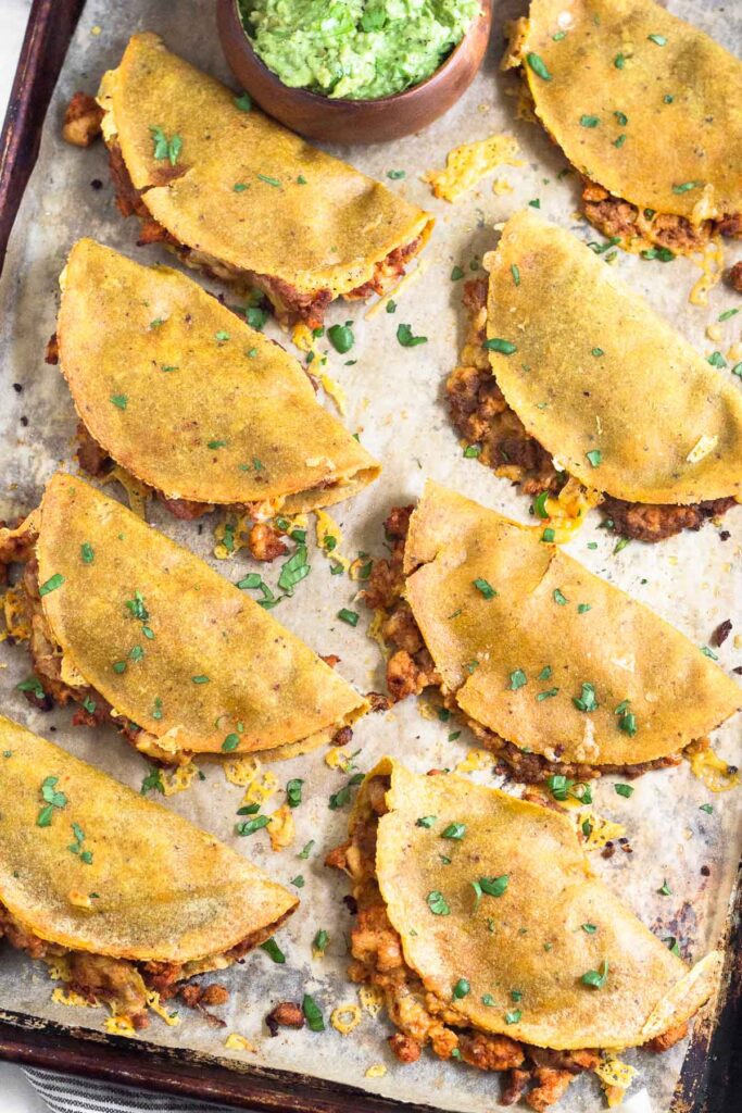 Overhead shot of oven baked tacos with some meat and cheese coming out of them. They are garnished with cilantro and there is a bowl of guacamole on the baking sheet.