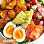 Breakfast bowl filled with greens, roasted potatoes, tomato and cucumber salad, smoked salmon, sliced avocado, and a hard-boiled egg and topped with everything but the bagel seasoning. Next to it is half an avocado.