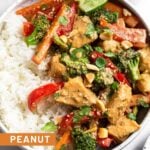 Peanut butter chicken in a bowl with white rice topped with cilantro, peanuts, sesame seeds and a lime wedge. Behind the bowl is a kitchen towel and a small bowl of peanuts.