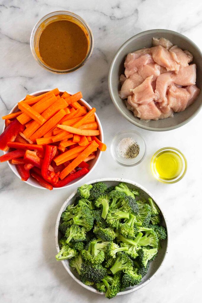 White marble counter top with a bowl of diced raw chicken, a bowl of olive oil, a bowl of salt and pepper, a bowl of broccoli florets, a bowl of julienned carrots and bell peppers, and a jar of peanut sauce.
