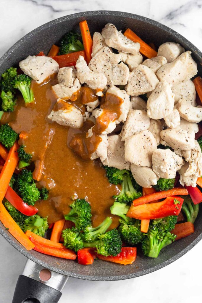 Large skillet filled with sautéed broccoli, carrots, red bell peppers, and sautéed bite-sized pieces of chicken with peanut sauce poured overtop.