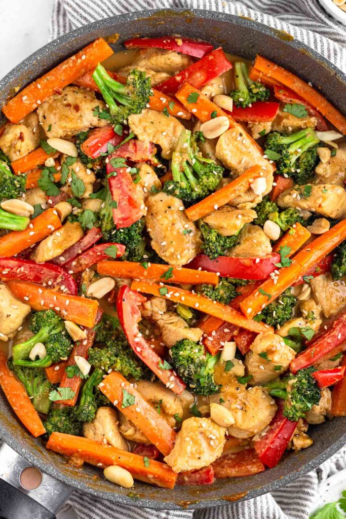 A large pan of chicken and veggie stir fry tossed with peanut sauce and topped with cilantro, sesame seeds, and peanuts.