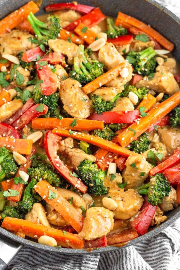Peanut chicken with veggies in a large skillet topped with peanuts, cilantro, and sesame seeds.