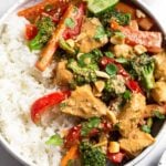 Peanut butter chicken in a bowl with white rice topped with cilantro, peanuts, sesame seeds and a lime wedge. Behind the bowl is a kitchen towel and a small bowl of peanuts.