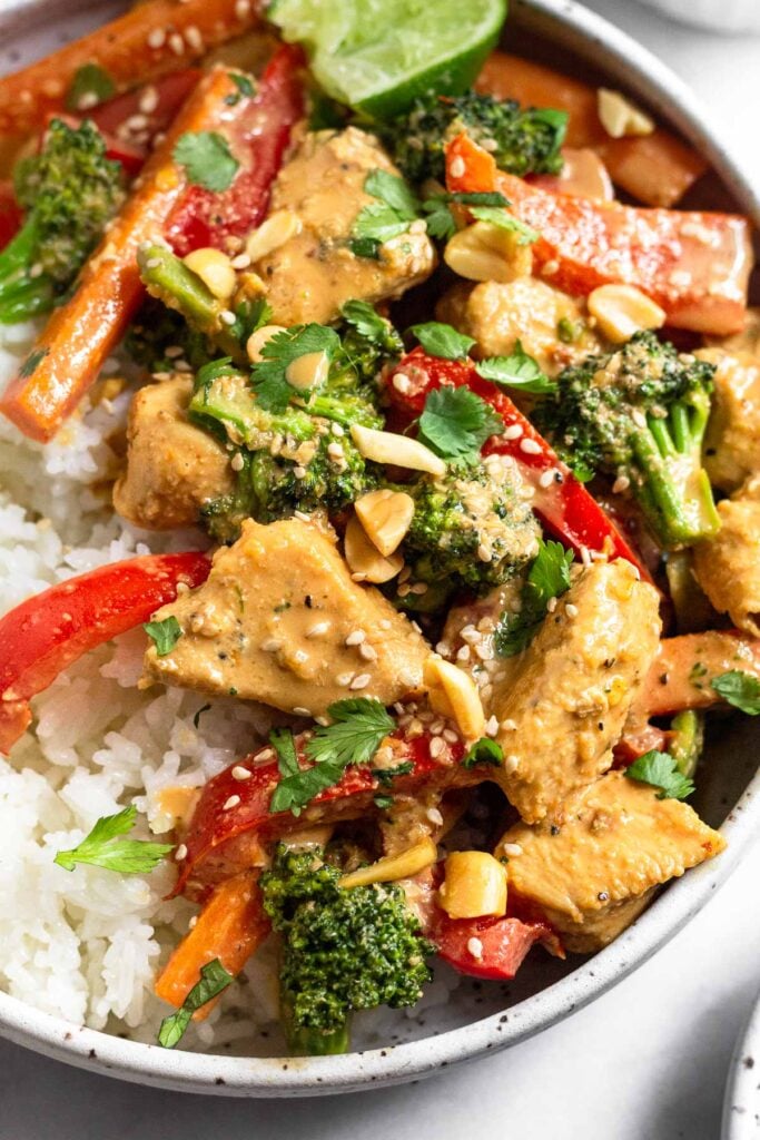 Thai peanut chicken and white rice in a bowl. It is garnished with cilantro, peanuts, sesame seeds and a lime wedge.
