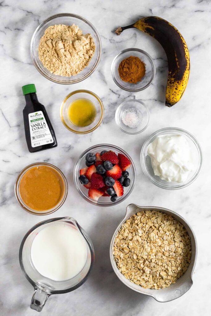 White marble counter with a bowl banana, a bowl of spices, a bowl of salt, a bowl of greek yogurt, a bowl of rolled oats, a glass of milk, a bowl of diced berries, a bowl of peanut butter, a container of vanilla extract, a small bowl of honey, and a bowl of protein powder on it.