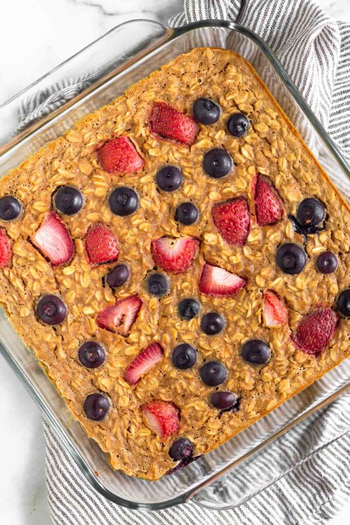 Overhead shot of protein baked oatmeal with strawberries and blueberries in a glass baking dish sitting on a kitchen towel.