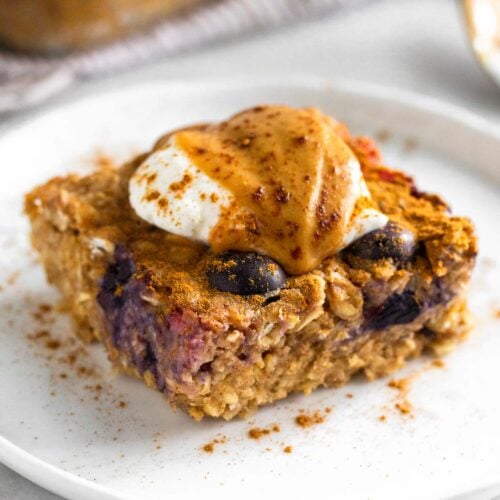 A piece of protein baked oatmeal on a plate topped with yogurt, peanut butter, and cinnamon. Behind it is the pan of the rest of the oatmeal.
