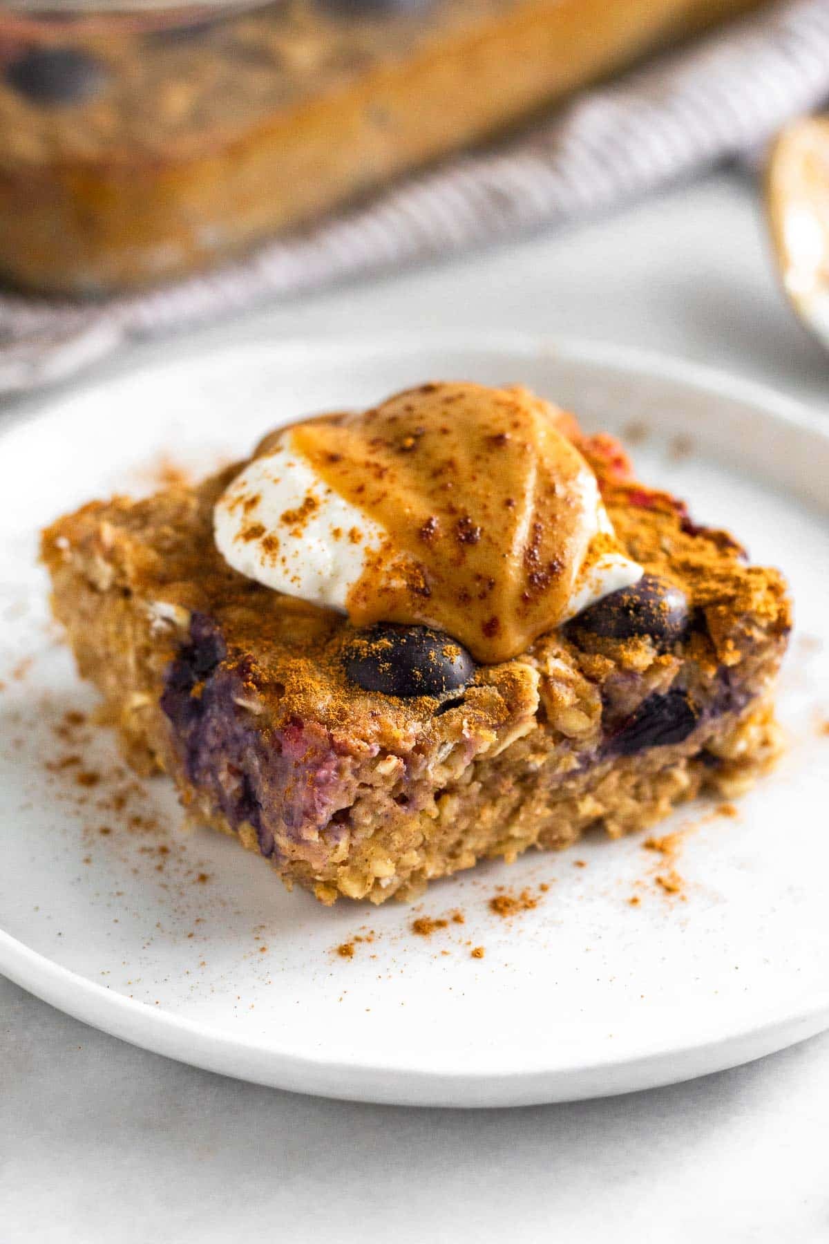 A piece of protein baked oatmeal on a plate topped with yogurt, peanut butter, and cinnamon. Behind it is the pan of the rest of the oatmeal.