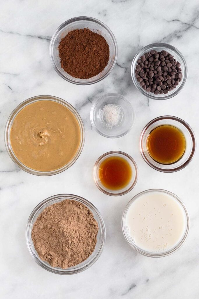 Marble counter of a bowl of cocoa powder, a bowl of chocolate chips, a bowl of maple syrup, a bowl of milk, a bowl of vanilla extract, a bowl of chocolate protein powder, a bowl of nut butter, and a bowl of salt.