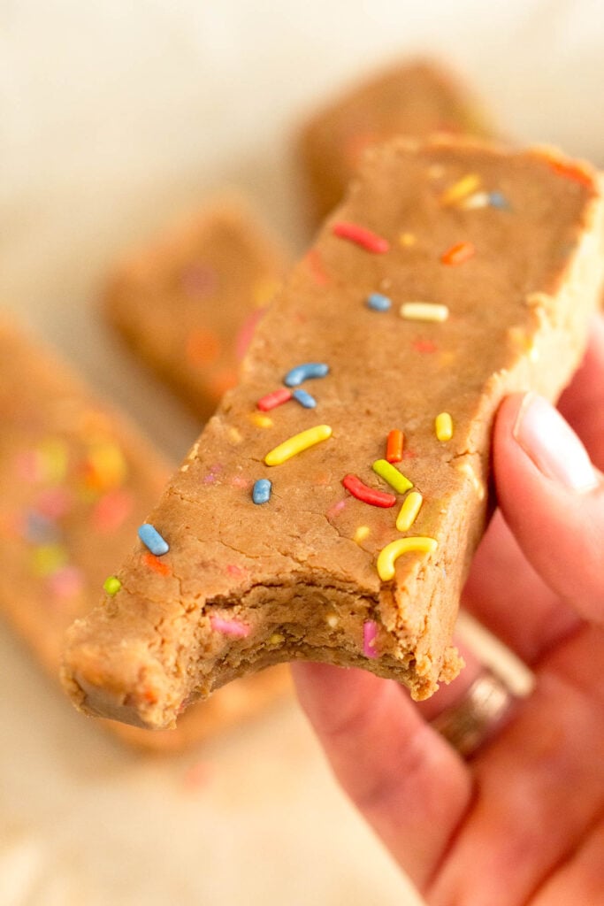 A hand holding a homemade protein bar with sprinkles in it with a bite taken out of it. In the background are more bars.