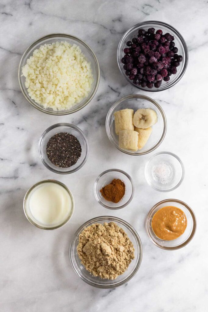 White marble counter with a bowl of frozen blueberries, a bowl of frozen banana pieces, a bowl of salt, a bowl of peanut butter, a bowl of protein powder, a bowl of cinnamon, a jar of milk, a bowl of chia seeds, and a bowl of frozen cauliflower rice.
