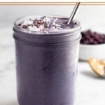 Blueberry hormone balancing smoothie in a jar topped with coconut shreds and cacao nibs with a straw in it. Behind it is a bowl of frozen blueberries and a spoon with peanut butter on it.