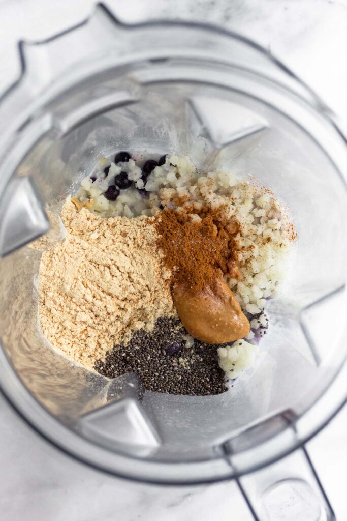 Blender filled with frozen fruit, frozen cauliflower rice, protein powder, chia seeds, peanut butter, cinnamon, and milk.