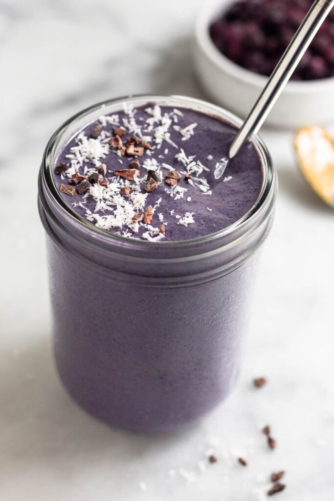 Healthy smoothie in a jar topped with coconut shreds and cacao nibs with a straw in it. Behind it is a bowl of frozen blueberries and a spoon with peanut butter on it.