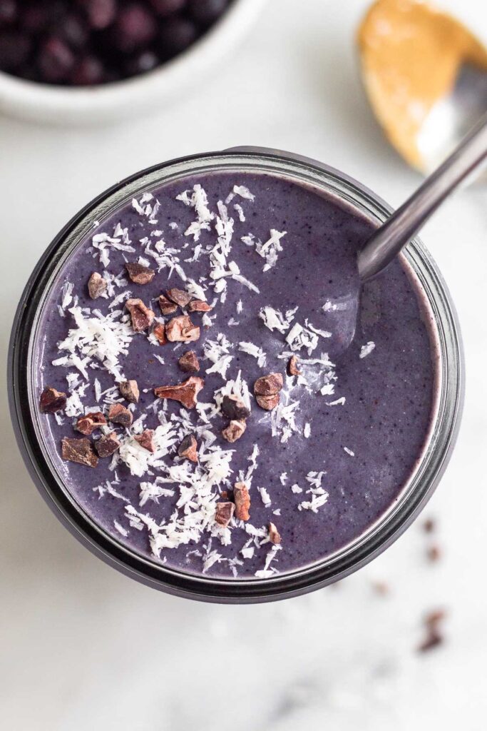 Overhead shot of a purple smoothie topped with shredded coconut and cacao nibs and a straw in it. Behind it is a bowl of frozen blueberries and a spoon with peanut butter on it.