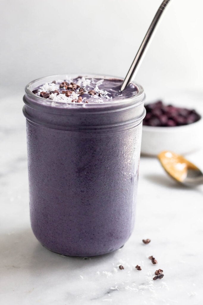 Blueberry hormone balancing smoothie in a jar topped with coconut shreds and cacao nibs with a straw in it. Behind it is a bowl of frozen blueberries and a spoon with peanut butter on it.
