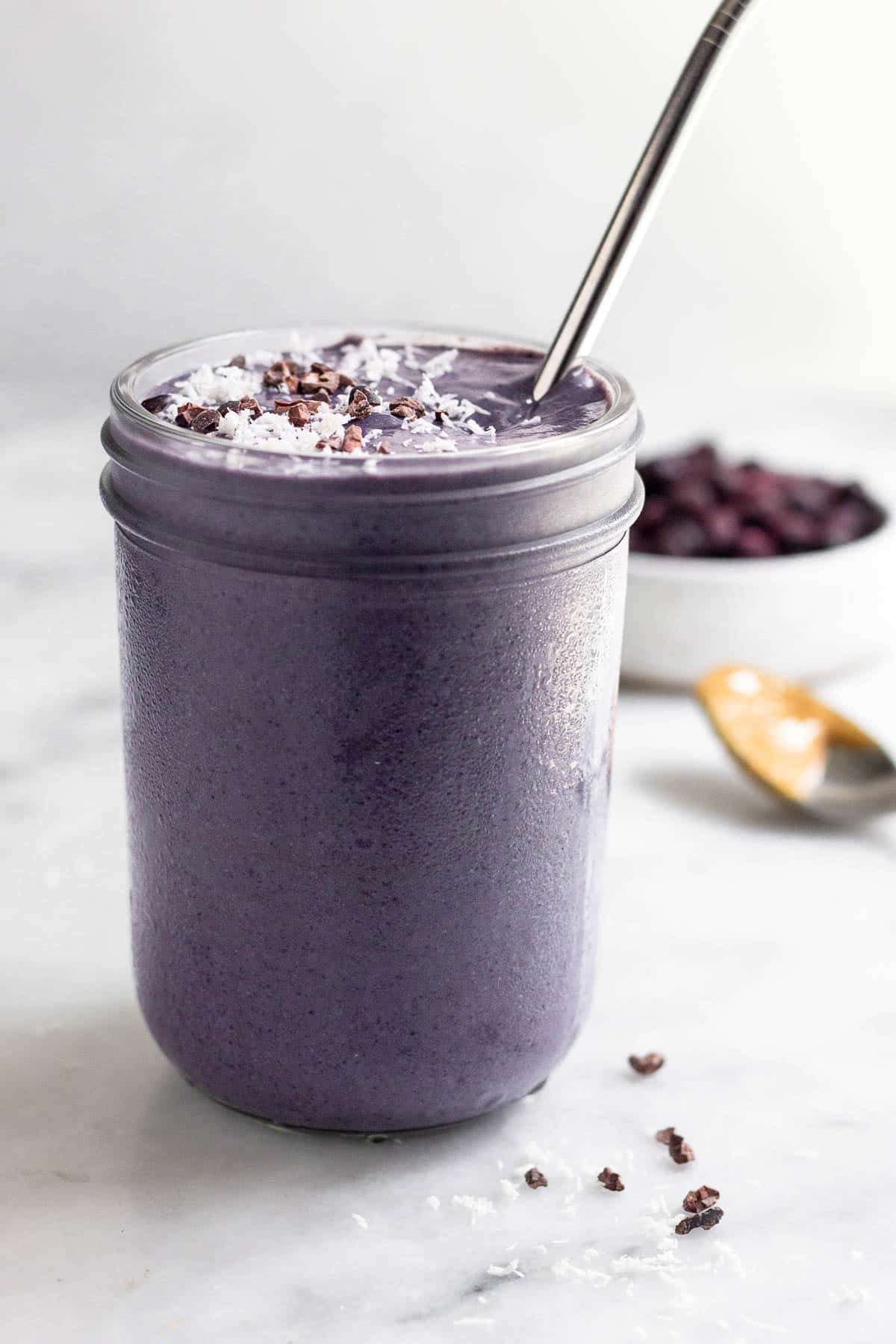 Blueberry hormone balancing smoothie in a jar topped with coconut shreds and cacao nibs with a straw in it. Behind it is a bowl of frozen blueberries and a spoon with peanut butter on it.