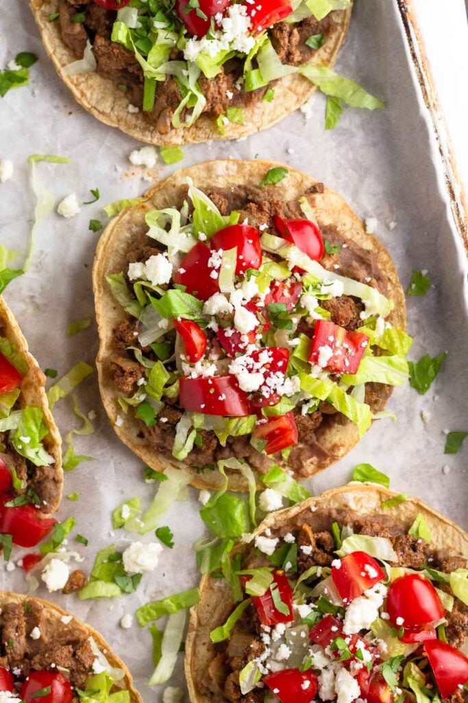 A baking sheet with ground beef tostadas with beans, taco meat, lettuce, tomato, cheese, and cilantro.