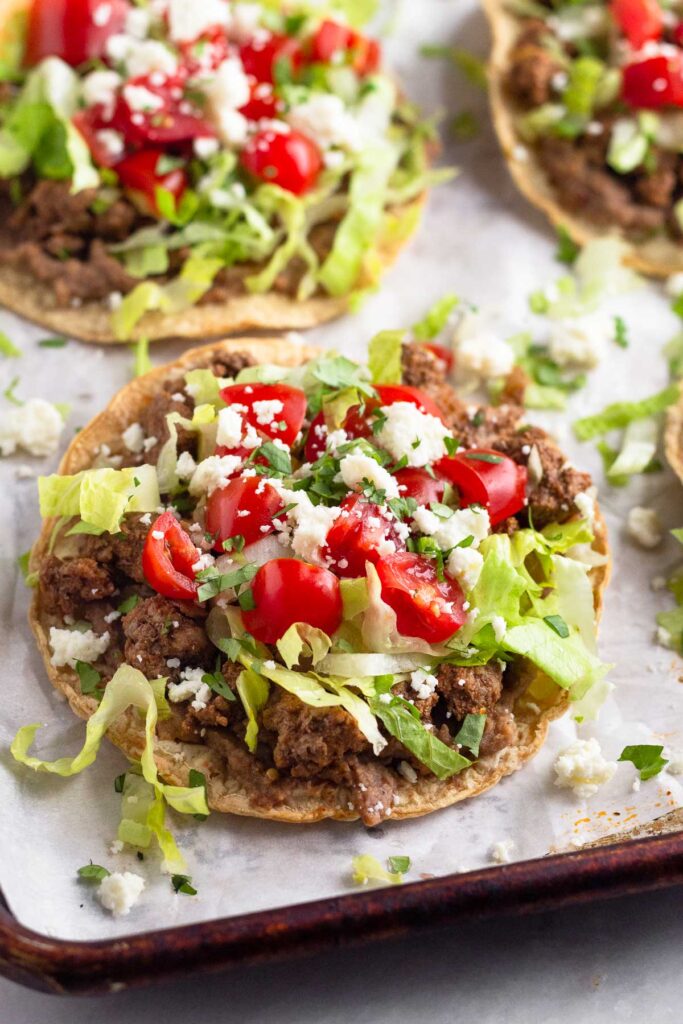 Beef tostadas with beans, lettuce, tomato, cheese, and cilantro on a baking sheet lined with parchment paper.