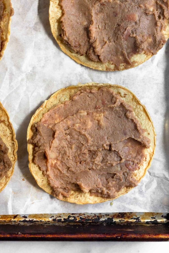 Crispy corn tortilla shell with refried beans spread across it on a baking sheet lined with parchment paper.