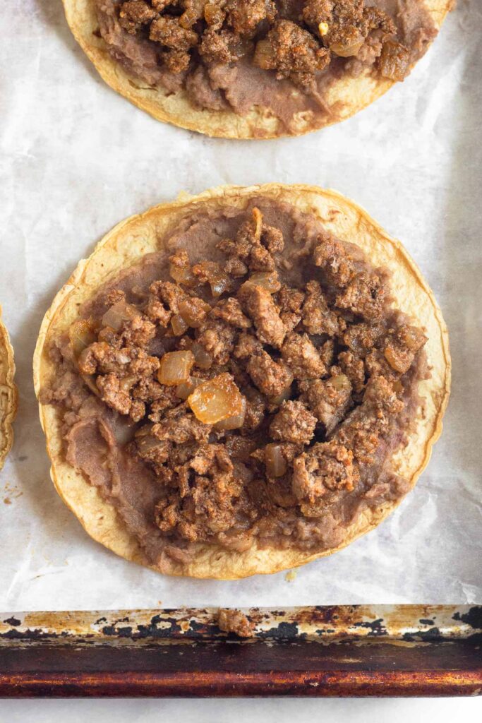 Crispy corn tortilla shell with refried beans spread across it. On top of the beans is taco meat. It is on a baking sheet lined with parchment paper.