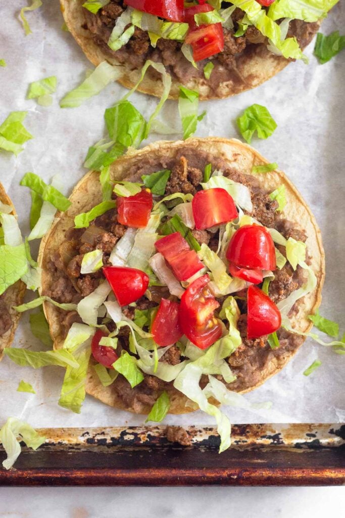 Beef and bean tostadas topped with lettuce and tomato on baking sheet.