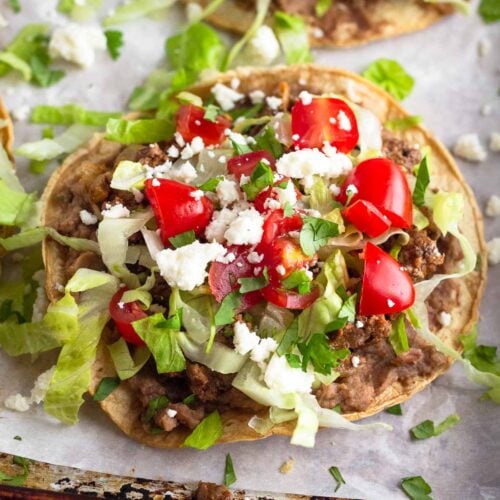 Beef tostada with beans, taco meat, lettuce, tomato, cheese, and cilantro. Behind it is another one and they are on a baking sheet lined with parchment.
