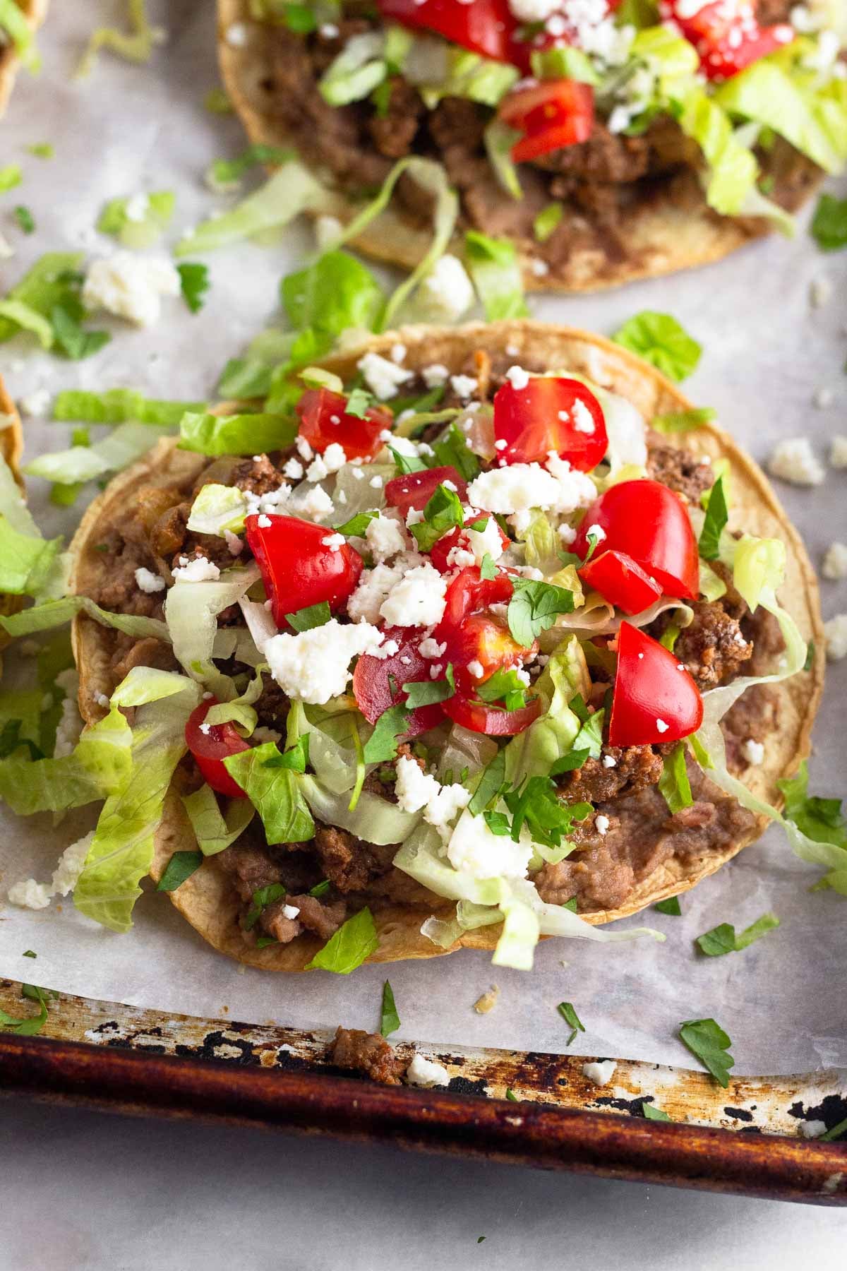 Beef tostada with beans, taco meat, lettuce, tomato, cheese, and cilantro. Behind it is another one and they are on a baking sheet lined with parchment.