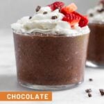 Chocolate chia pudding in a small glass topped with whipped cream, strawberries, and cacao nibs. Behind it is a similar glass and around them is cacao nibs.