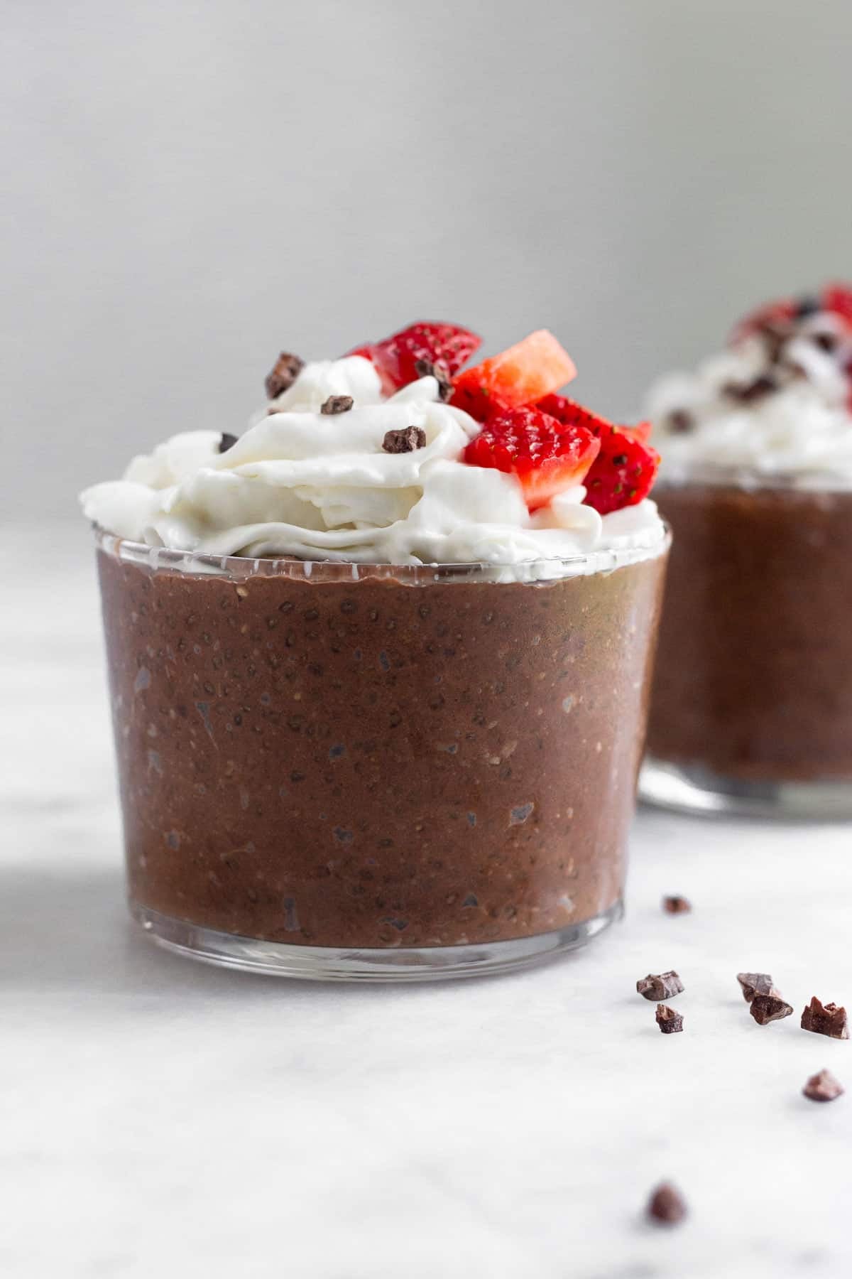 Chocolate chia pudding in a small glass topped with whipped cream, strawberries, and cacao nibs. Behind it is a similar glass and around them is cacao nibs.