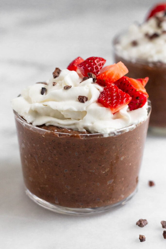 Chocolate chia seed pudding in a small glass topped with whipped cream, strawberries, and cacao nibs. Behind it is a similar glass.