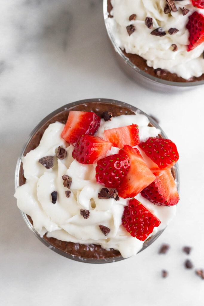 Overhead shot of chocolate coconut chia pudding in a small jar with whipped cream, strawberries, and cacao nibs on top of it. Off to the side is another jar with the same thing.