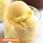 Scoops of pineapple dole whip in a small glass cup. Behind it is another cup and a bowl of frozen pineapple.