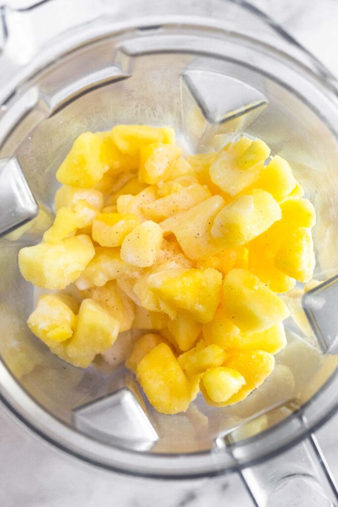 An overtop shot of a blender filled with frozen pineapple chunks, coconut milk, and salt.
