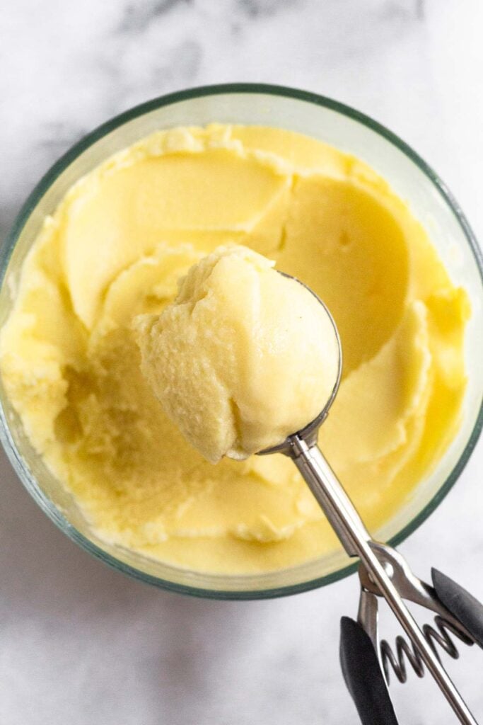 Glass bowl filled with pineapple dole whip. Overtop of it is an ice cream scoop filled with it.