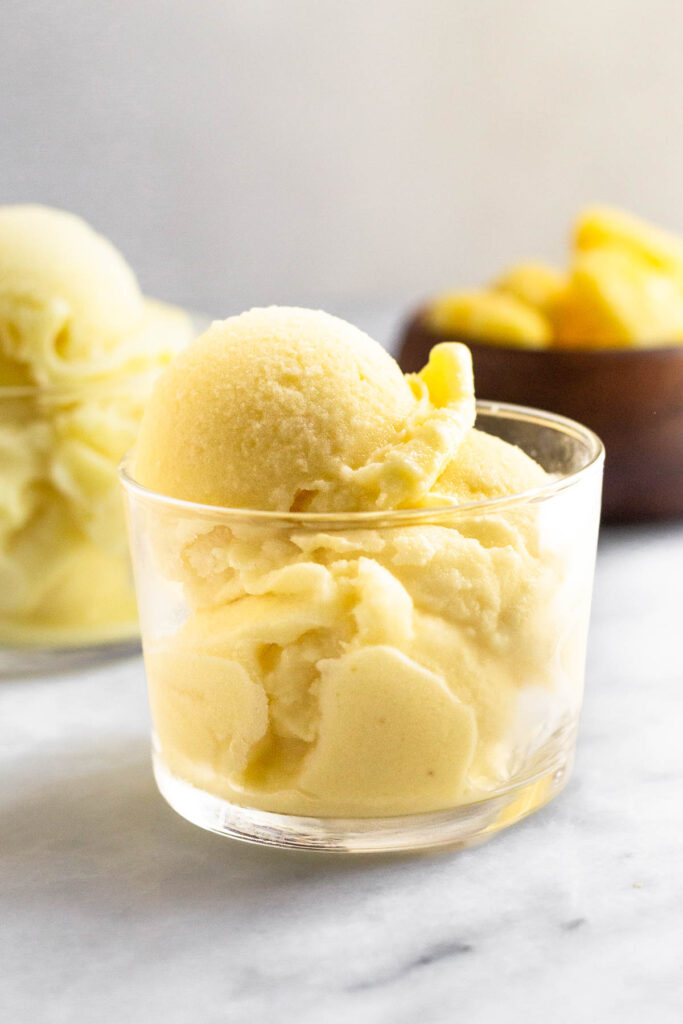 Homemade dole whip in a small glass. Behind it is another filled glass and a bowl of frozen pineapple.